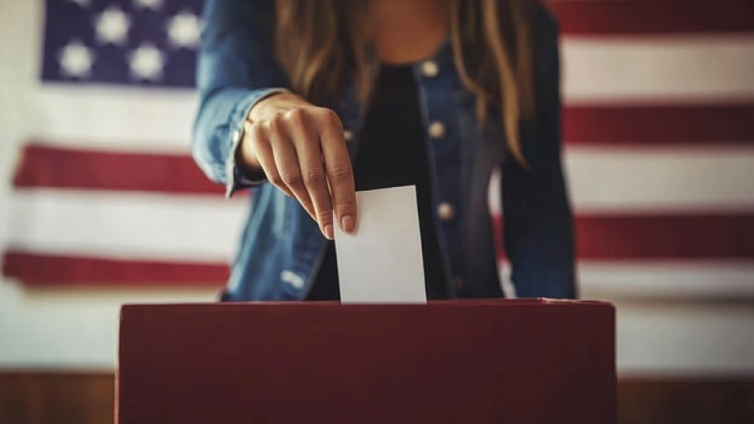 Female putting voting ballot into ballot box with US flag behind her